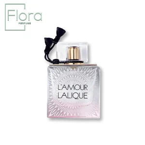 تصویری از شیشه عطر لالیک لامور را نشان میدهد. این شیشه به شکل مربع و با طراحی شفاف همراه با نقشهای ظریف و گلبرگهای سفید بر روی بدنه دیده میشود. درب شیشه به رنگ طلایی و شفاف است و توسط بندی تزیینی در کنار آن آراسته شده است. نام “L’AMOUR LALIQUE” با فونتی مشکی و برجسته بر روی شیشه نوشته شده است. در سمت بالا چپ تصویر، لوگوی “Flora PERFUME” به چشم میخورد.