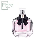 تصویری از یک بطری عطر ایو سن لورن مون پاریس (YSL Mon Paris) با طراحی شیشهای شفاف و محتوی مایع صورتیرنگ. گردن بطری با یک پاپیون مشکی زیبا و آویز طلایی با لوگوی YSL تزئین شده است. لوگوی Flora Perfume در گوشه بالا، سمت چپ تصویر مشاهده میشود.